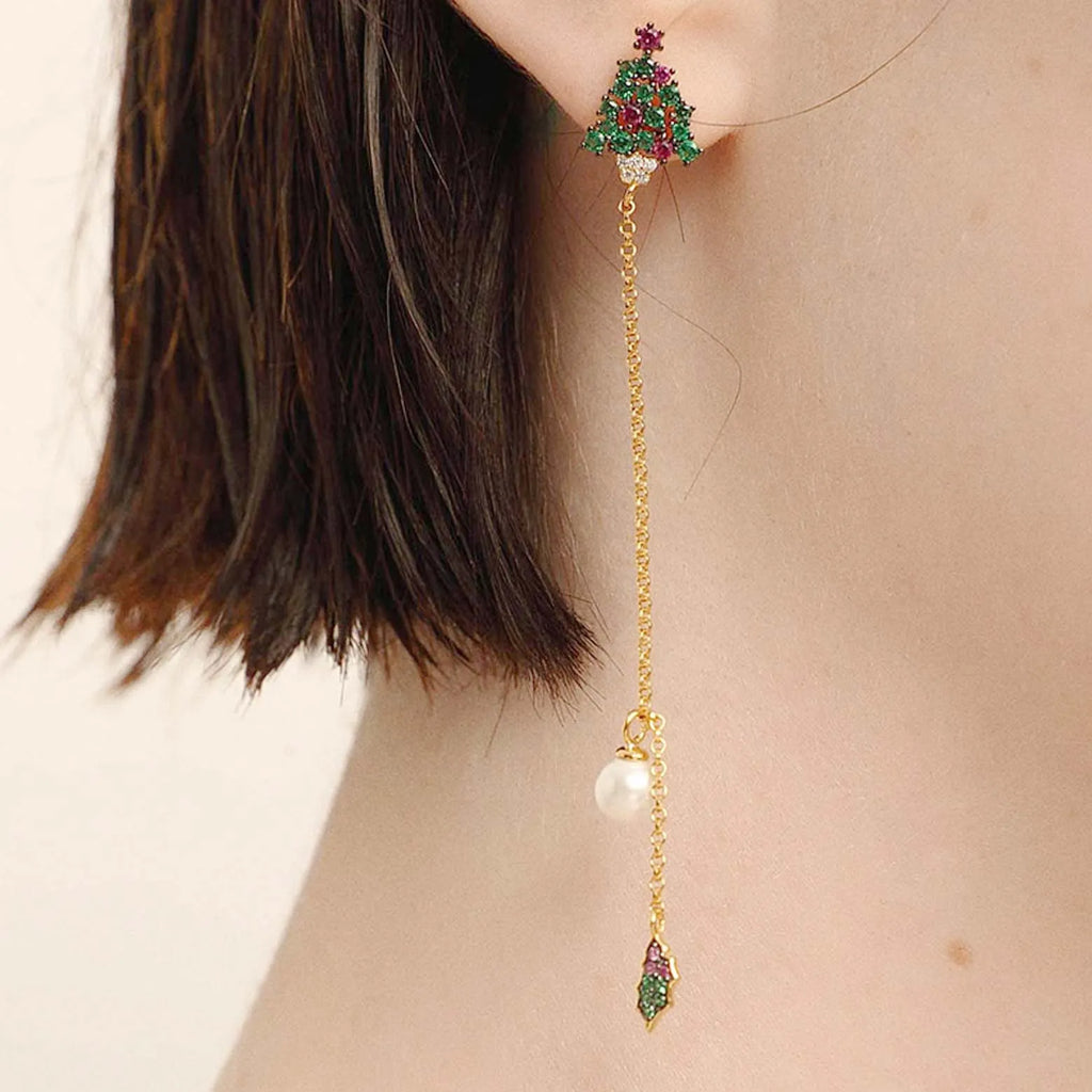 A side view of a woman wearing a unique, long gold earring. The earring begins with a green gemstone Christmas tree-shaped stud, detailed with multicolored gem ornaments, connected by a fine chain to a single white pearl, creating a festive and elegant holiday accessory.