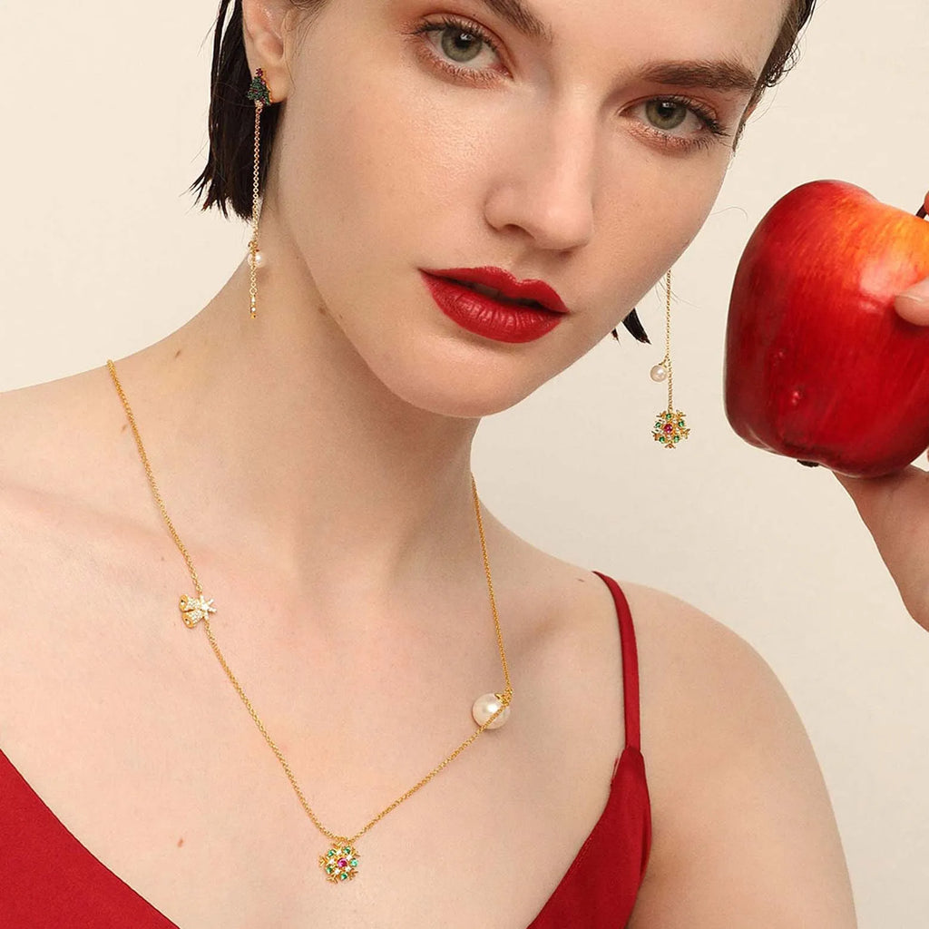 A woman in a red top, holding a red apple close to her face, wears a fine gold necklace with a butterfly charm and a sunburst pendant encrusted with multicolored gemstones, with a single pearl on the chain. She also has matching gold earrings with a chain link, a small green gemstone, and a dangling pearl.