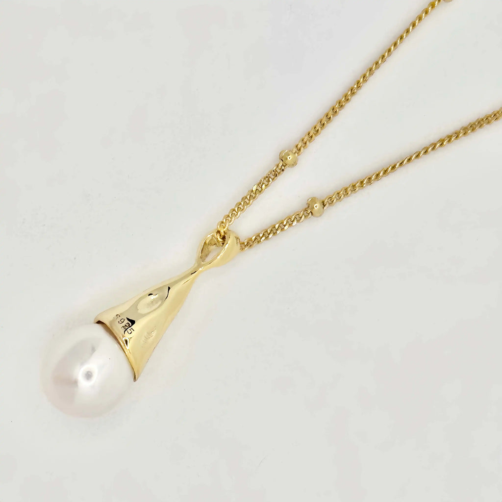 The image displays a close-up of a gold pendant earring with a large, round, white pearl. The pearl is set in a gold cap which connects to a delicate gold chain. The gold has a polished finish and is marked with "925," indicating that the metal is sterling silver that has been gold-plated. The pearl exhibits a soft luster and a smooth surface, reflecting the light in a gentle glow. The background is a soft, neutral color, which complements the warmth of the gold and the serene beauty of the pearl.