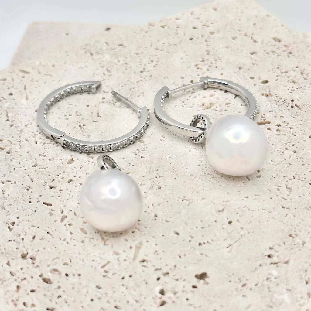 This image shows a pair of silver hoop earrings, each lined with small, clear crystals that provide a sparkling accent. Below the hoops, there are two large, white pearls with a notable sheen, reflecting light and showing hints of iridescence. The pearls are detached from the hoops, suggesting they may be interchangeable or part of a set. The earrings and pearls rest against a textured, beige surface that resembles sandstone, allowing the jewelry's polished finish to stand out.