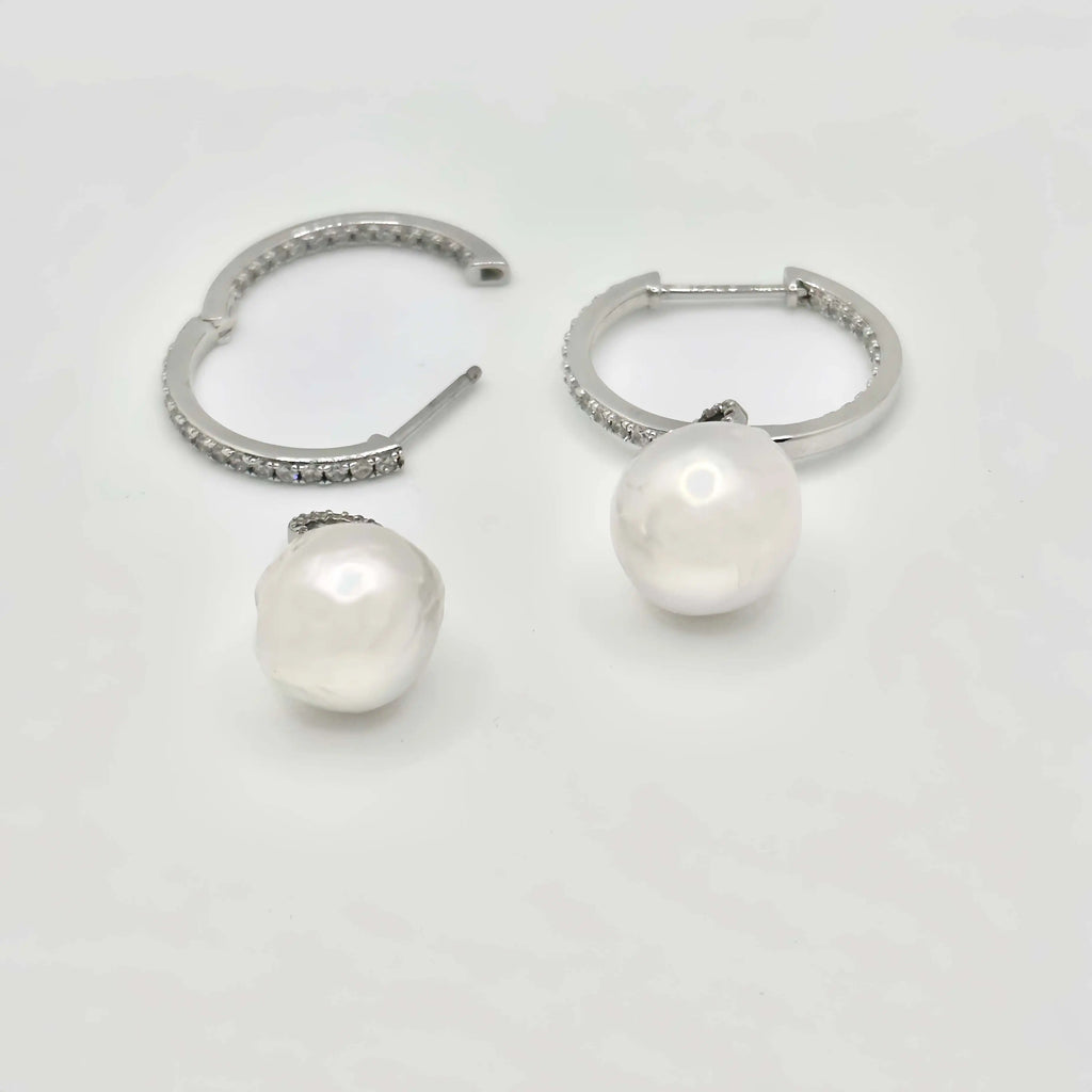 This image shows a pair of silver hoop earrings, each lined with small, clear crystals that provide a sparkling accent. Below the hoops, there are two large, white pearls with a notable sheen, reflecting light and showing hints of iridescence. The pearls are detached from the hoops, suggesting they may be interchangeable or part of a set. The earrings and pearls rest against a textured, beige surface that resembles sandstone, allowing the jewelry's polished finish to stand out.