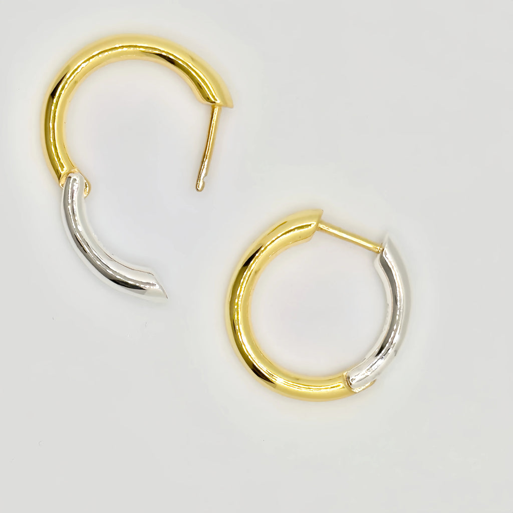 A pair of modern gold and silver hoop earrings, featuring a two-tone design with one half of each hoop in polished gold and the other in silver. The earrings have a sleek, open hoop style with a post closure, lying against a white background.
