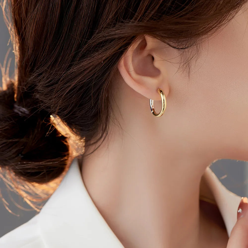 A woman is shown from the side, gently holding a small gold hoop earring with her fingers, preparing to adorn her ear with its mate, which is already in place. The earrings are simple yet elegant, reflecting the light and complementing her sophisticated, minimalistic style.