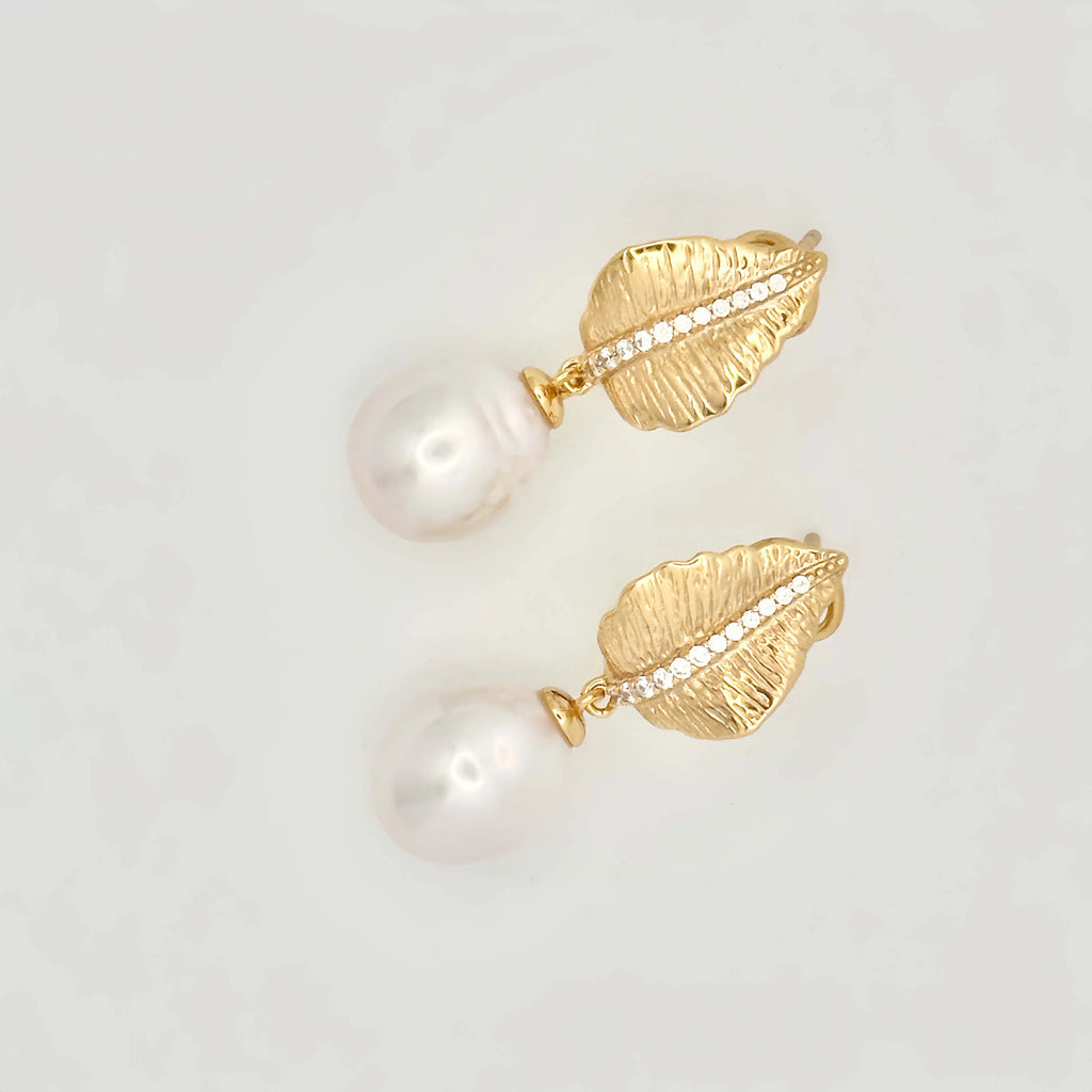 A pair of sophisticated earrings with a nature-inspired design, featuring large white pearls. Each pearl is mounted on a gold setting that resembles a textured leaf or petal, accented with a line of small diamonds that add sparkle to the organic motif, all set against a neutral background.