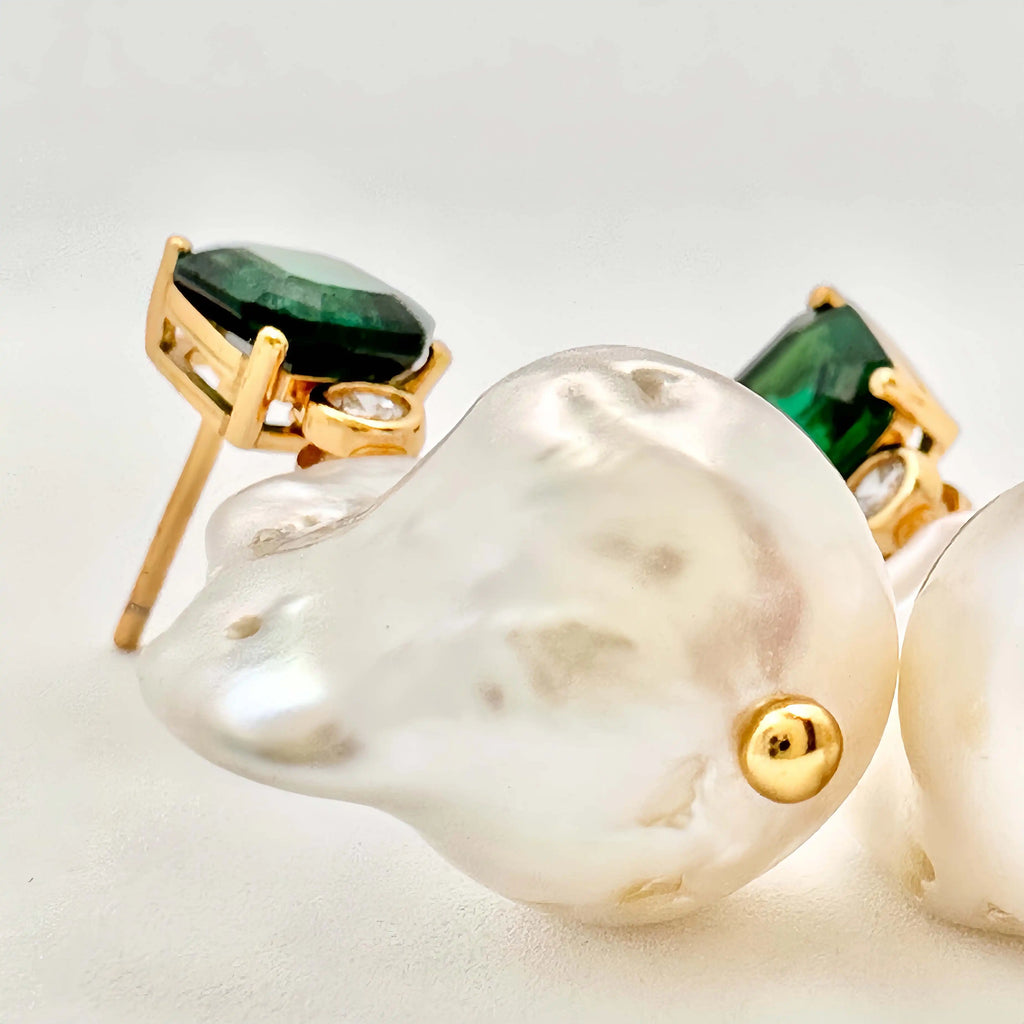 A pair of elegant earrings presented on a white background. Each earring features a large square-cut green gemstone set in gold, with a small round diamond below it, leading to an irregularly shaped baroque pearl that adds a touch of organic sophistication to the design.