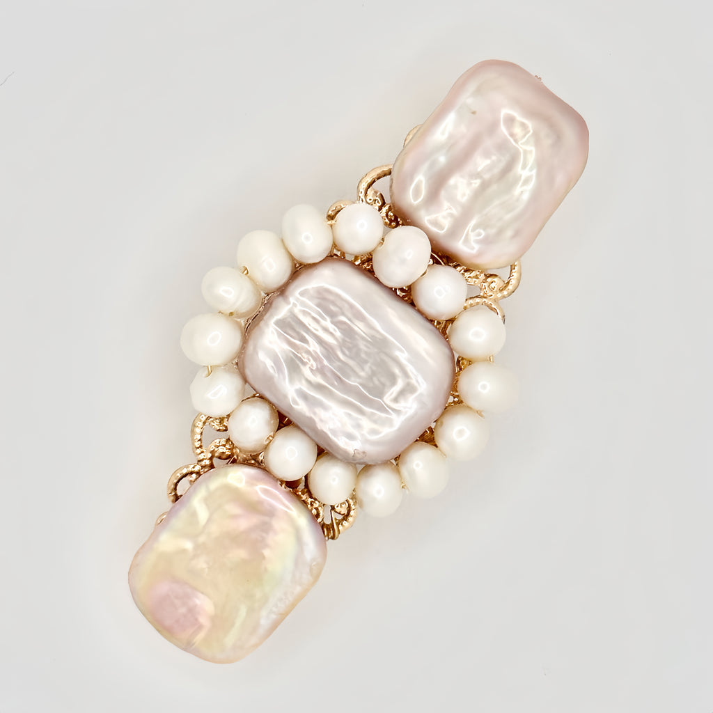 The image shows a gold-toned hair clip adorned with a central rectangular pink pearl, flanked by two irregular-shaped pearls with a lustrous sheen reflecting pastel hues. Surrounding the central pearls is a ring of smaller, round white pearls, set against the intricate gold filigree detailing of the clip. The piece exudes a blend of classic elegance and modern charm, set against a neutral background that highlights its colors and textures.