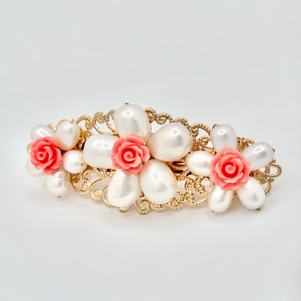  The image displays a golden hair clip featuring a trio of white pearl flowers, each with a delicate pink rose at the center. The pearls have an irregular, natural shape, giving the flowers a lifelike appearance. The gold setting has a filigree design that adds a vintage touch to the accessory. The background is a clean, solid color, putting the focus on the intricate details of the hair clip.