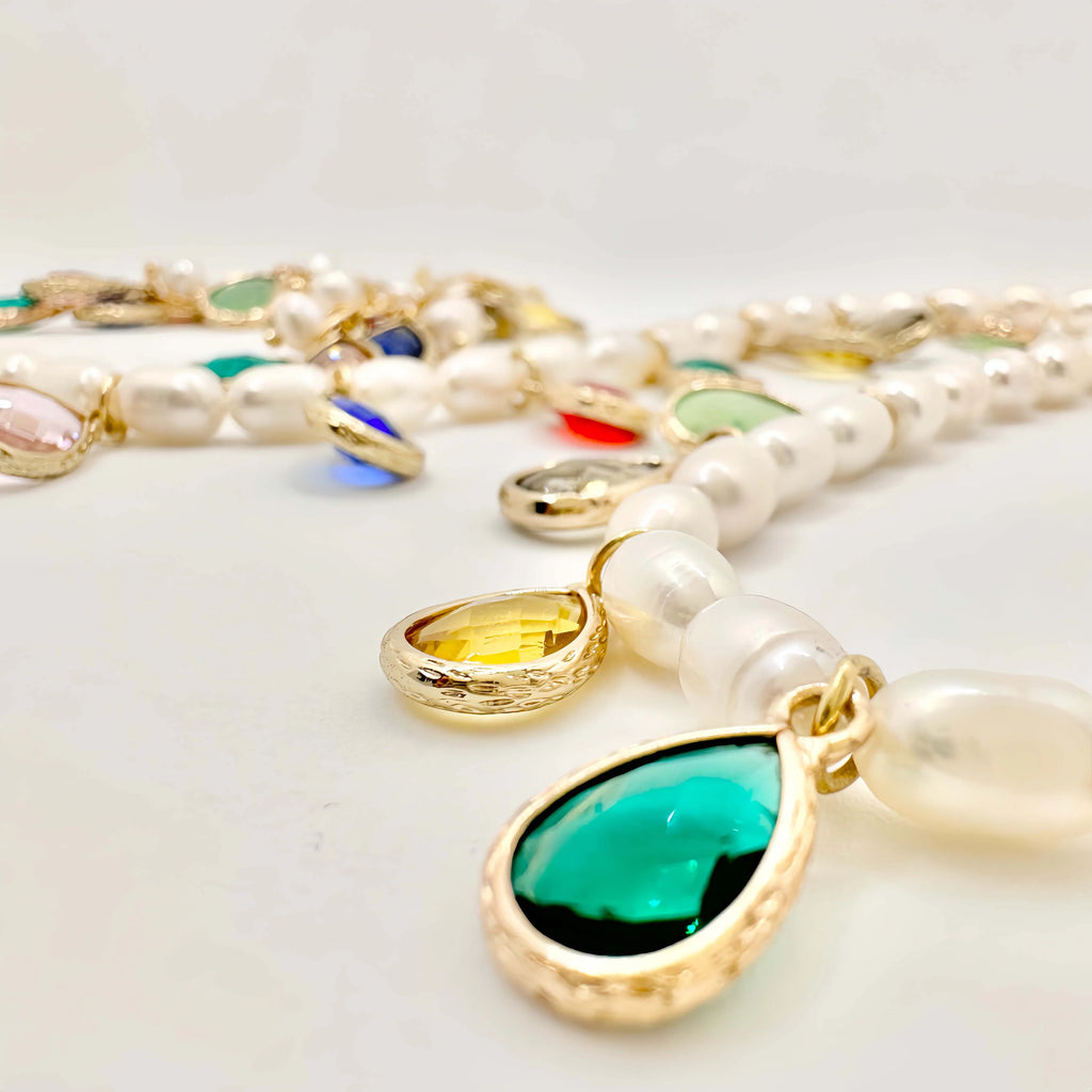 A luxurious pearl necklace and bracelet set displayed on a white background. The necklace is a string of round white pearls, interspersed with gold links and teardrop-shaped colored gemstones in bezel settings. The bracelet mirrors the design with similar colored gems and smaller pearls, both creating a harmonious and opulent look.