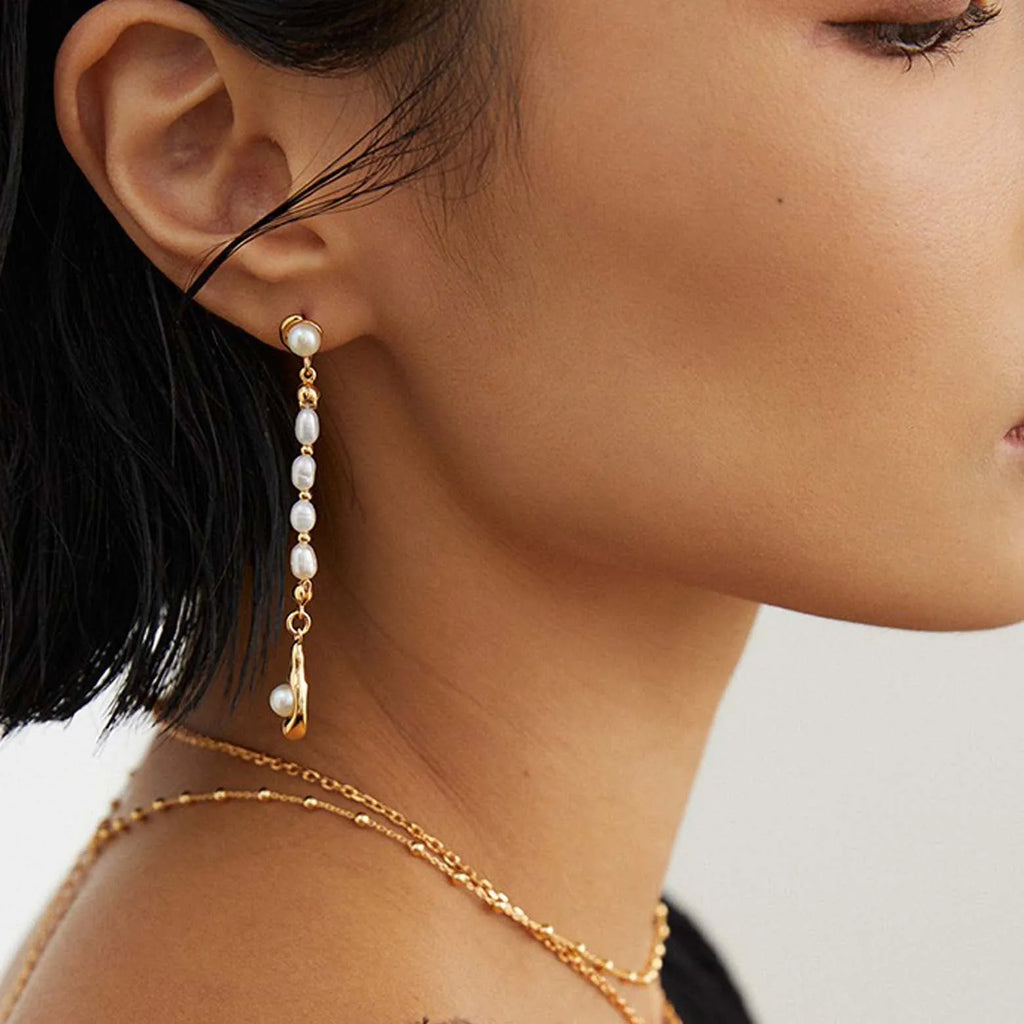 The image depicts a close-up side profile of a woman wearing a long, elegant gold earring. The earring features a single large pearl at the lobe and a vertical chain of smaller pearls descending in a linear fashion. The pearls vary slightly in size and have a rich luster, with the entire piece exuding a luxurious and contemporary feel. The woman's hand, adorned with a gold ring and her nails painted in a neutral shade with an accent design, is gently touching the earring, drawing attention to it.