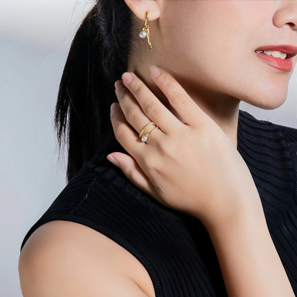 A close-up side view of a woman with a low ponytail, wearing a sophisticated black pleated dress with a high collar. She is adorned with a delicate gold earring that has a small pearl and a crystal detail, giving a subtle hint of elegance to her overall attire.