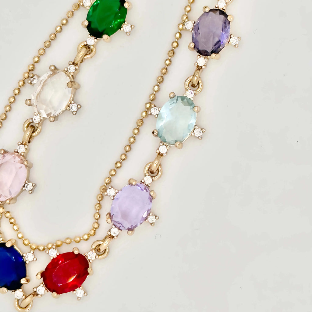 A striking gold necklace laid out on a white surface, featuring a chain of diverse, multicolored gemstones, each set in a gold frame. The necklace transitions into a simple gold chain that culminates in a large, baroque pearl pendant, creating an elegant drop effect. The piece is a blend of classic and contemporary styles.