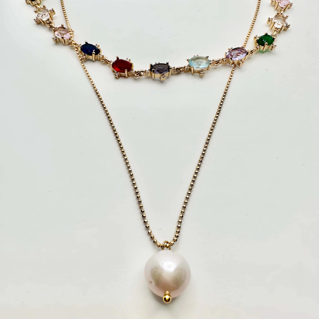 A striking gold necklace laid out on a white surface, featuring a chain of diverse, multicolored gemstones, each set in a gold frame. The necklace transitions into a simple gold chain that culminates in a large, baroque pearl pendant, creating an elegant drop effect. The piece is a blend of classic and contemporary styles.