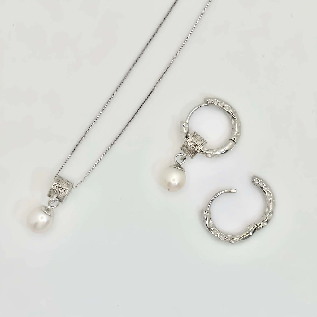 A pair of silver earrings displayed on a white background. Each earring features a textured hoop with an organic, molten-like design, attached to a decorative square bead with intricate patterns. A classic white pearl dangles elegantly from the bead, adding a touch of timeless elegance to the modern design.