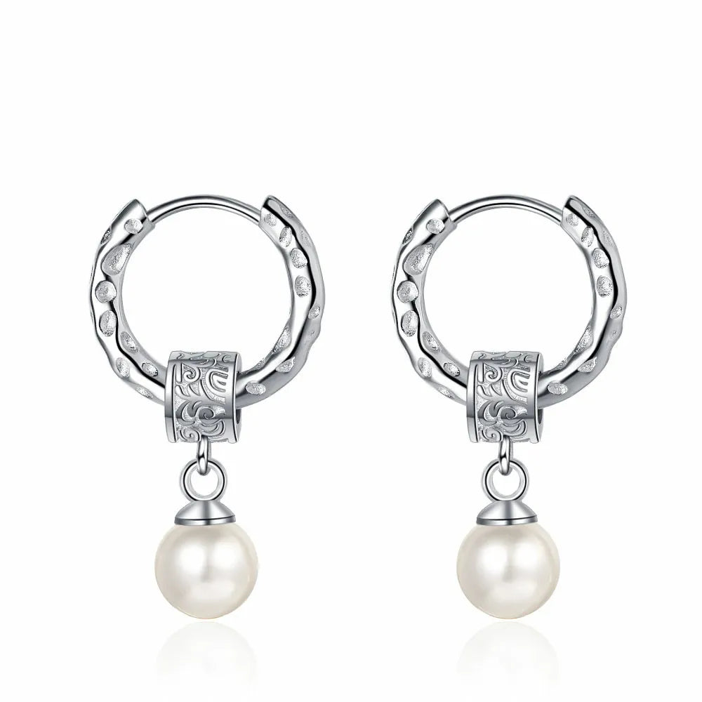 A pair of silver earrings displayed on a white background. Each earring features a textured hoop with an organic, molten-like design, attached to a decorative square bead with intricate patterns. A classic white pearl dangles elegantly from the bead, adding a touch of timeless elegance to the modern design.