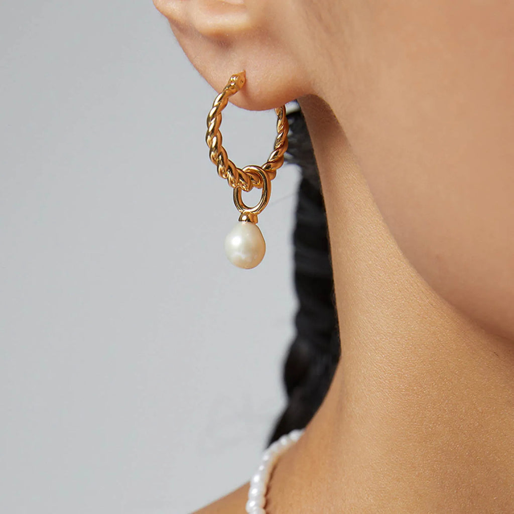 This image features a woman's profile, highlighting a silver hoop earring with a twisted design worn on her ear. A single, luminous pearl dangles from the hoop, providing a classic and elegant touch. The pearl's iridescent surface captures the light, contrasting with the metallic sheen of the earring. The woman's hair is pulled back to showcase the earring fully, and her attire appears to be a simple white top or dress, which complements the understated sophistication of the jewelry.