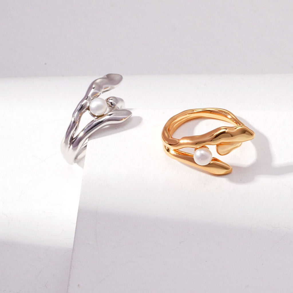 This image presents a gold ring with a unique and fluid design, resembling gentle waves that wrap around the finger. At the crest of one of the waves sits a small, round, white pearl that adds an element of classic elegance to the otherwise modern design. The gold has a polished, reflective surface that gleams brightly. The ring is showcased against a white background with an angular shadow, which creates a contrast that highlights the ring's dynamic curves and the pearl's soft luster.