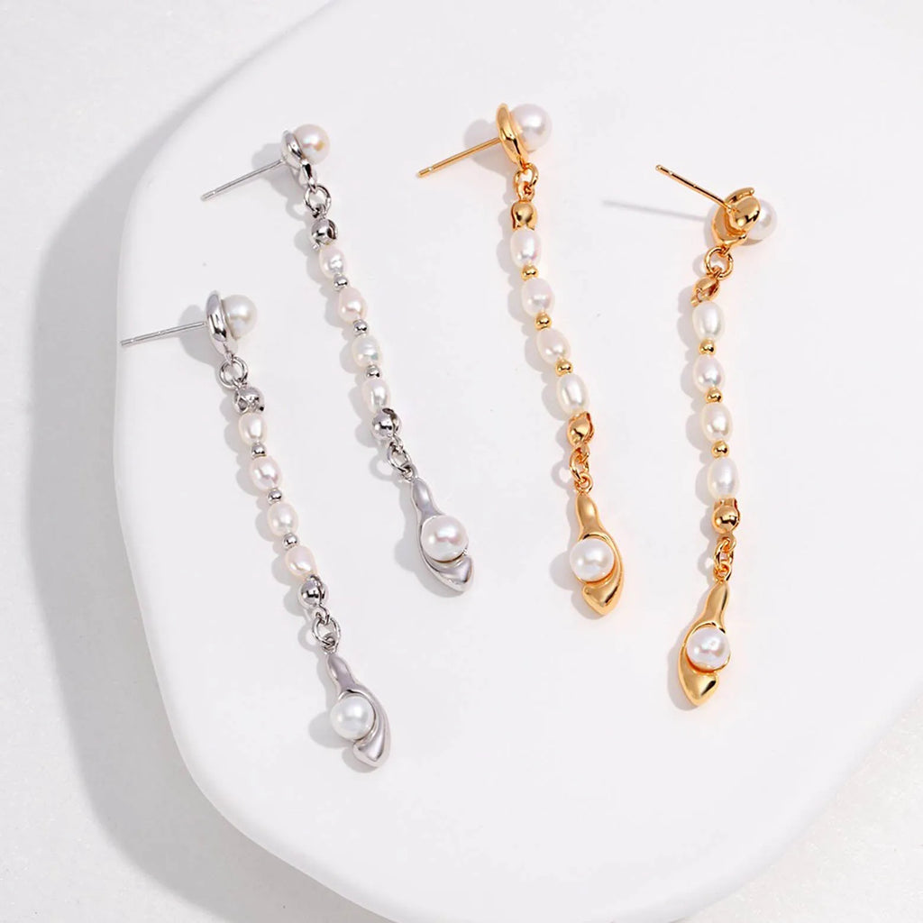 The image presents two pairs of long, elegant earrings, one in silver and the other in gold. Each earring features a series of small, round pearls in a vertical arrangement, connected by delicate metal links. The pearls are lustrous, with a gentle sheen that captures the light. At the bottom of each earring, a larger, unique pearl is cradled within a metal charm that echoes the shape of a flame or abstract figure. 