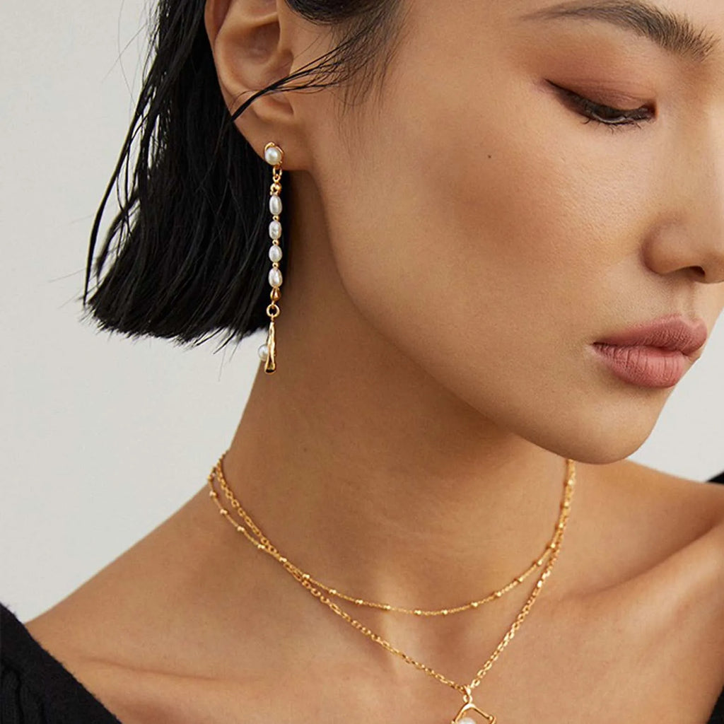 The image depicts a close-up side profile of a woman wearing a long, elegant gold earring. The earring features a single large pearl at the lobe and a vertical chain of smaller pearls descending in a linear fashion. The pearls vary slightly in size and have a rich luster, with the entire piece exuding a luxurious and contemporary feel. The woman's hand, adorned with a gold ring and her nails painted in a neutral shade with an accent design, is gently touching the earring, drawing attention to it.