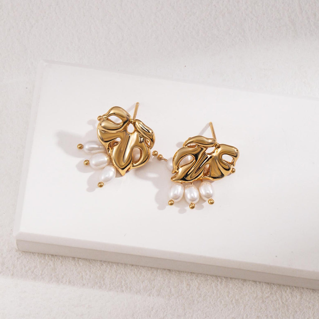 The image shows a pair of gold stud earrings, each featuring an abstract, openwork heart-shaped design. The hearts have a polished, sculptural quality, with smooth curves and holes that create a dynamic pattern. Suspended from each heart are three white oval pearls, attached by small gold links that end in round gold beads. 