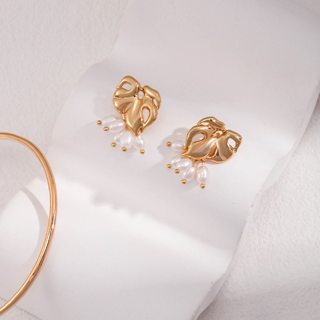 The image shows a pair of gold stud earrings, each featuring an abstract, openwork heart-shaped design. The hearts have a polished, sculptural quality, with smooth curves and holes that create a dynamic pattern. Suspended from each heart are three white oval pearls, attached by small gold links that end in round gold beads. 